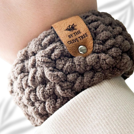 Brown faux suede label with by the olive tree logo attached to a dark brown crochet wristlet with a silver rivet fastener.